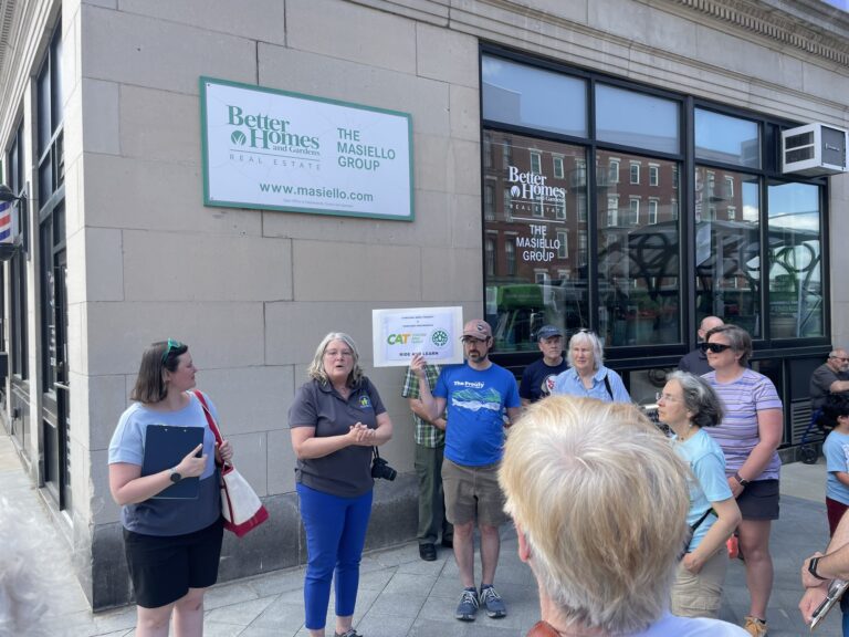 A woman in a dark gray shirt and blue pants greets a small gathering of people beside a gray downtown office building.